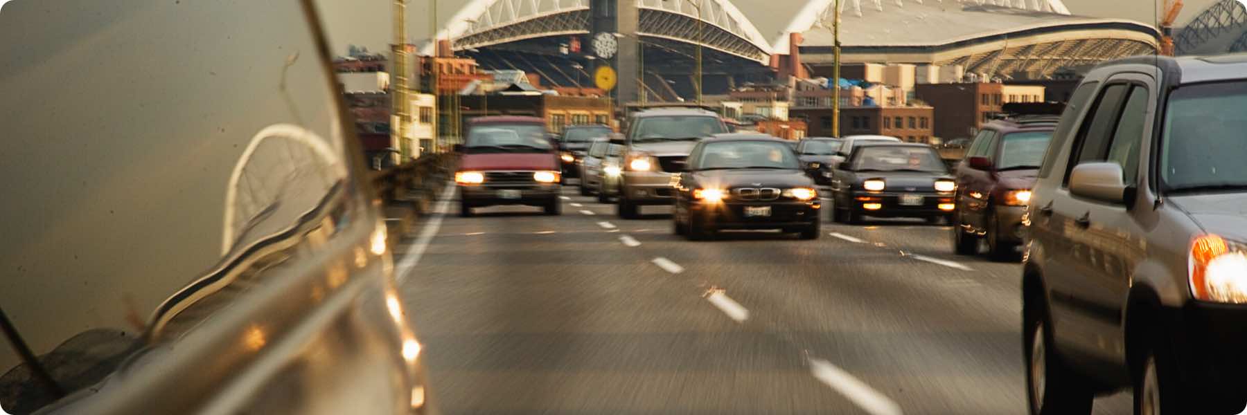 A busy multi-lane highway with cars streaming towards the camera sets the scene. The road, like a sleek web design, mirrors surrounding structures, while a large stadium looms in the background. This urban tableau reflects the hustle of a digital marketing agency amid city buildings.