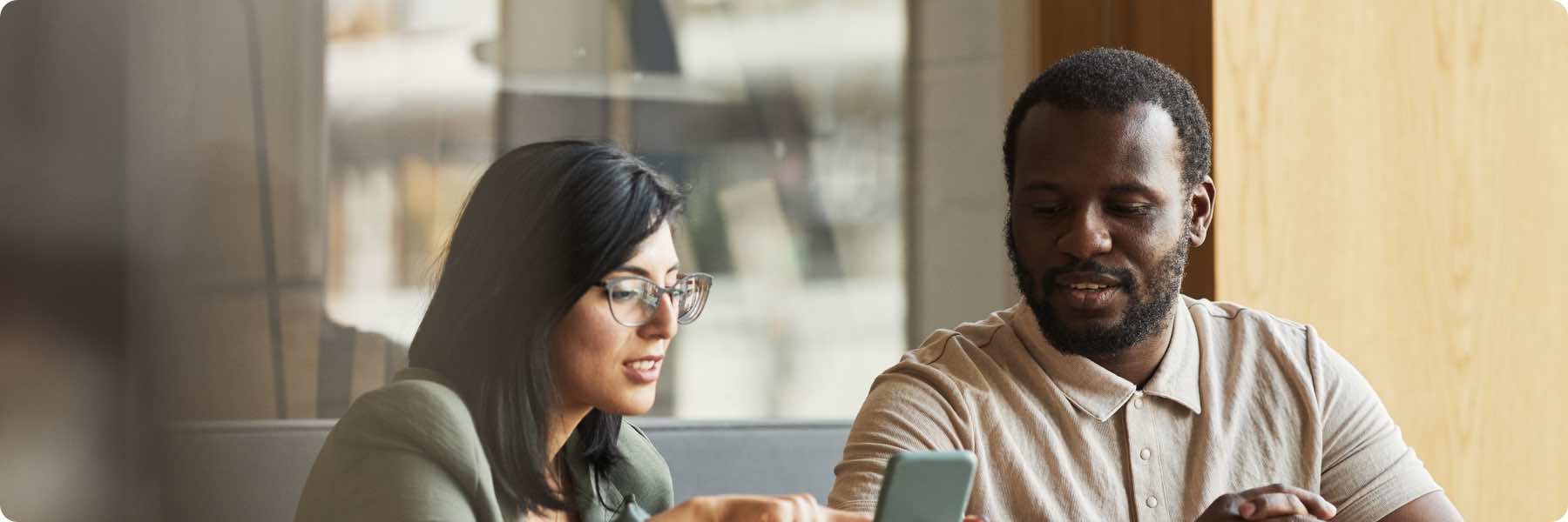 A man and woman sit together at a table, looking at a smartphone. The woman, wearing glasses, gestures towards the phone while discussing web design, as the man listens attentively. They appear to be in a casual indoor setting.