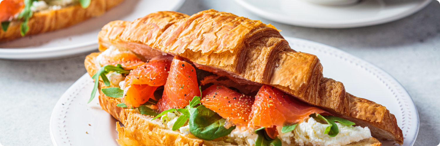 A croissant sandwich on a white plate, filled with smoked salmon, cream cheese, and greens, is ready to fuel your creativity like a marketing agency fuels innovation. Another similar plate is partially visible in the background, along with a cup on a saucer.
