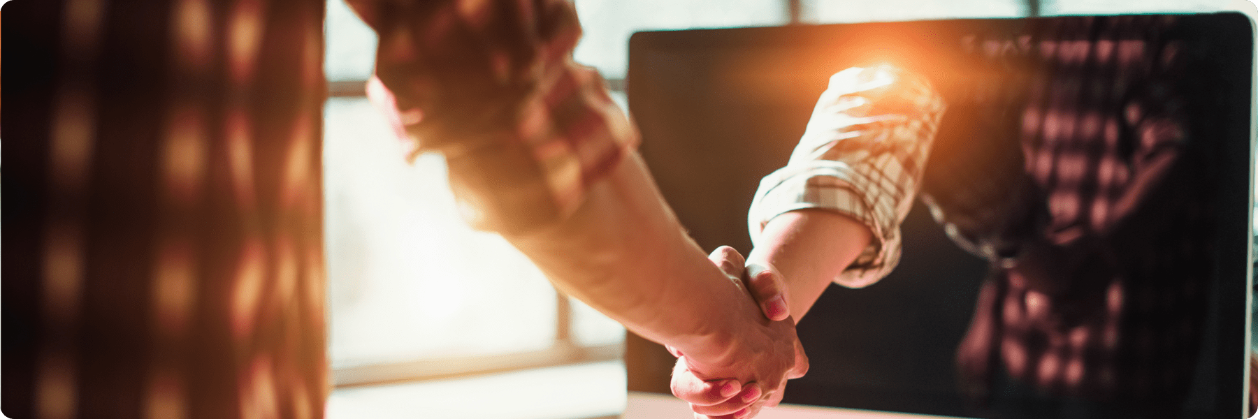 Two people shaking hands in front of a large monitor, forging a partnership with a digital marketing company. One person wears a plaid shirt, the other is visible only by their hand and sleeve. Warm lighting and a blurred background create a welcoming atmosphere.