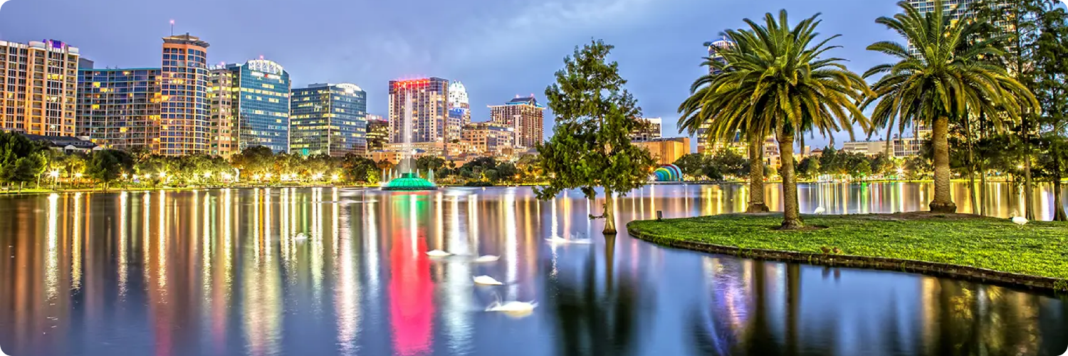 A vibrant cityscape at dusk with a shimmering lake in the foreground, much like a digital marketing agency illuminating your brand. High-rise buildings reflect colorful lights on the water, while palm trees and swans grace a small grassy area by the lake.