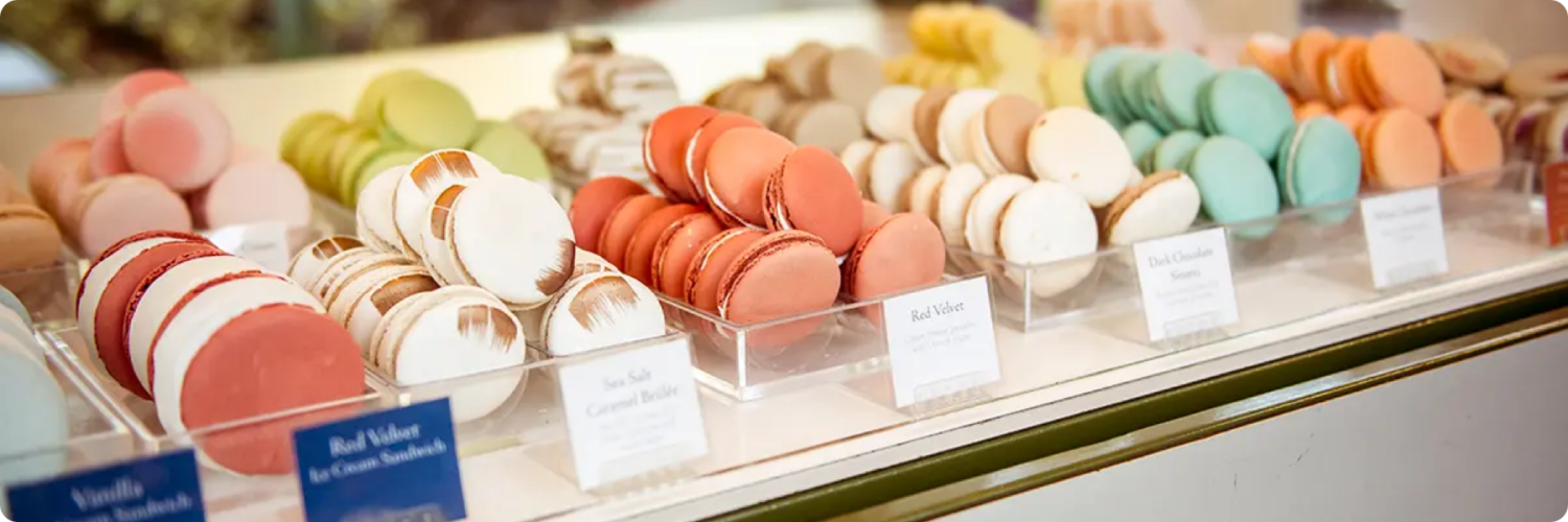 A display case filled with colorful macarons in various flavors, including red, pink, green, and blue. Each row features a label indicating the flavor, like Red Velvet and Smores, arranged neatly on transparent trays—crafted with the precision youd expect from a digital marketing company.