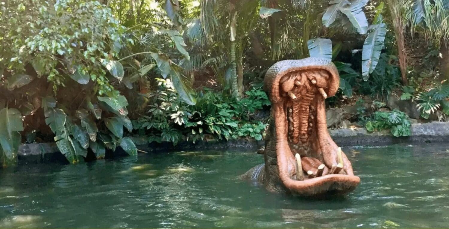 A large, realistic hippopotamus statue emerges from water with its mouth wide open, surrounded by dense, lush greenery and trees. The backdrop of tropical plants creates a jungle-like atmosphere—much like navigating the wilds of digital marketing for any leading agency.