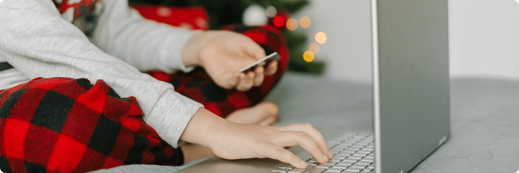 A child in plaid pants sits on a bed using a laptop, likely navigating a web design site, while holding a credit card. A Christmas tree with lights is blurred in the background, enhancing the festive atmosphere.
