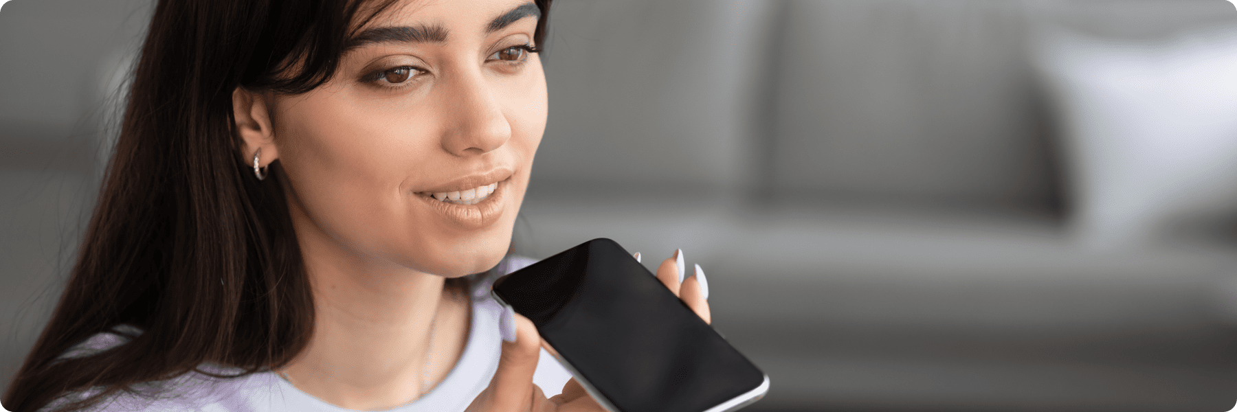 A woman, possibly from a digital marketing agency, holds a smartphone close to her mouth, appearing to speak into it. She is indoors with a blurred background, wearing a light-colored top and small hoop earrings, with long dark hair framing her face.