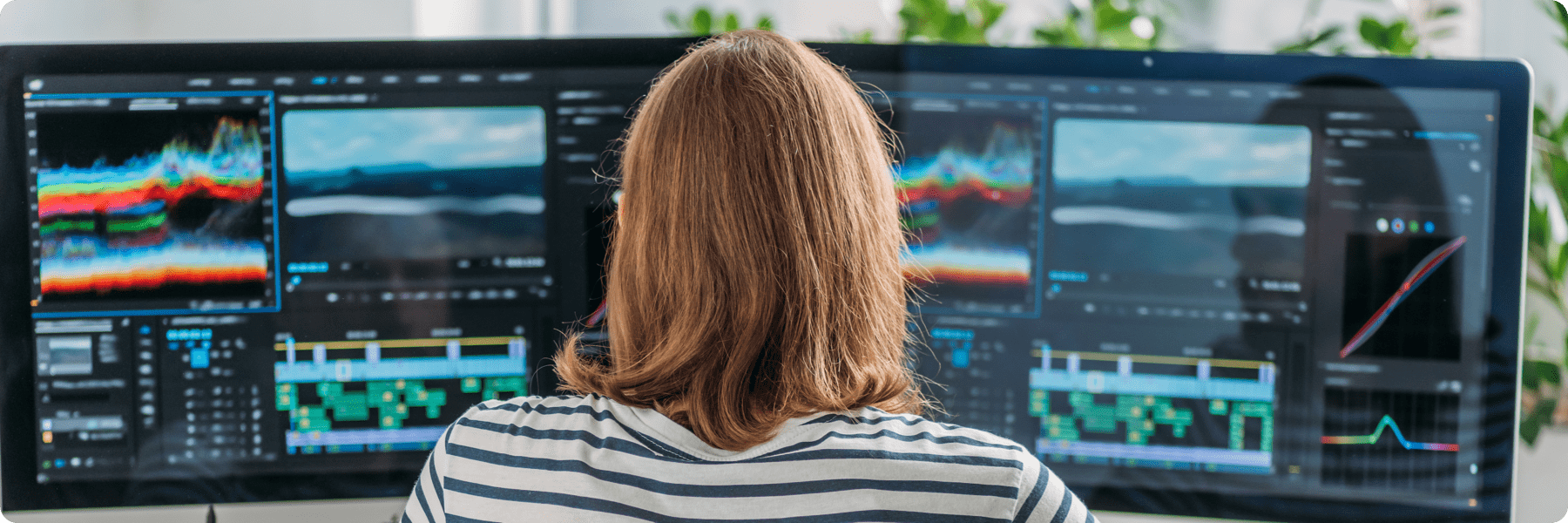 A person with shoulder-length hair, wearing a striped shirt, is working at a desk with multiple monitors displaying video editing software. The screens show colorful waveforms, timelines, and elements of a website project for a digital marketing company.