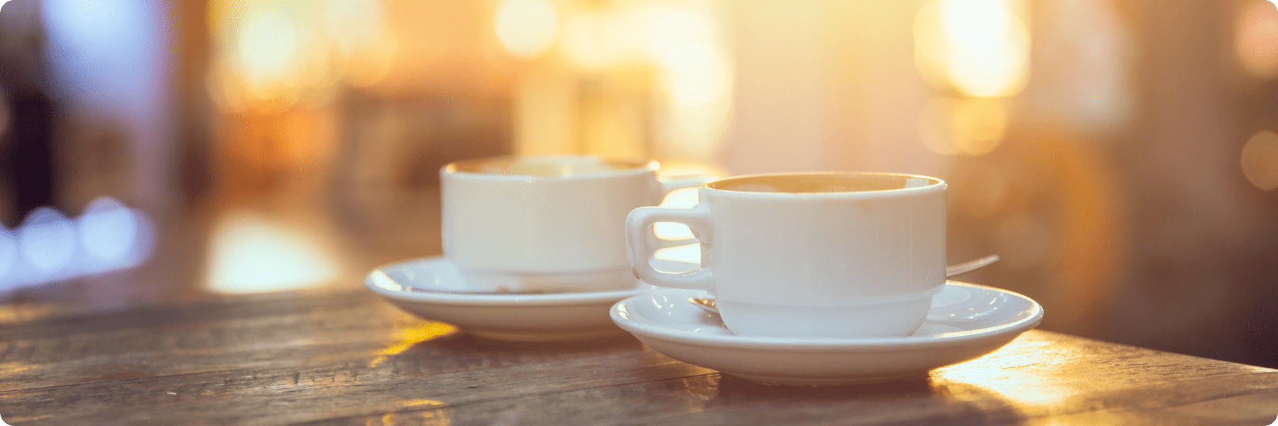Two white coffee cups with saucers sit on a wooden table, creating a cozy atmosphere reminiscent of a warm, welcoming digital marketing agency. The softly blurred background glows with golden light.