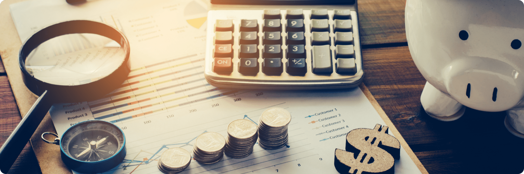 A wooden desk showcases financial tools: a magnifying glass, calculator, stack of coins, dollar sign, compass, and piggy bank, all arranged on a graph-laden chart. This scene subtly mirrors the meticulous planning at a digital marketing company.