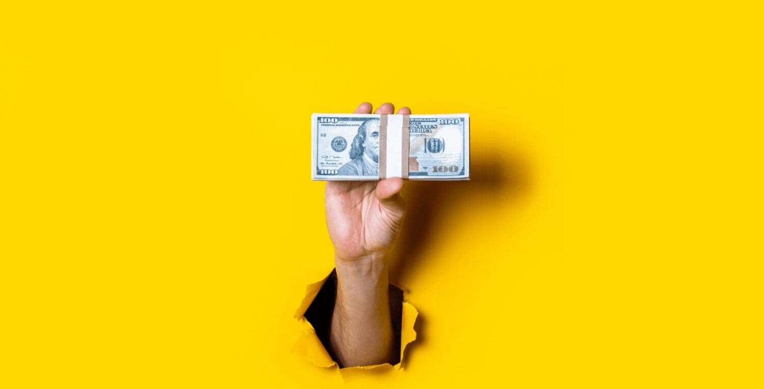 A hand from a digital marketing agency emerges through a tear in a bright yellow background, holding a one hundred dollar bill vertically.
