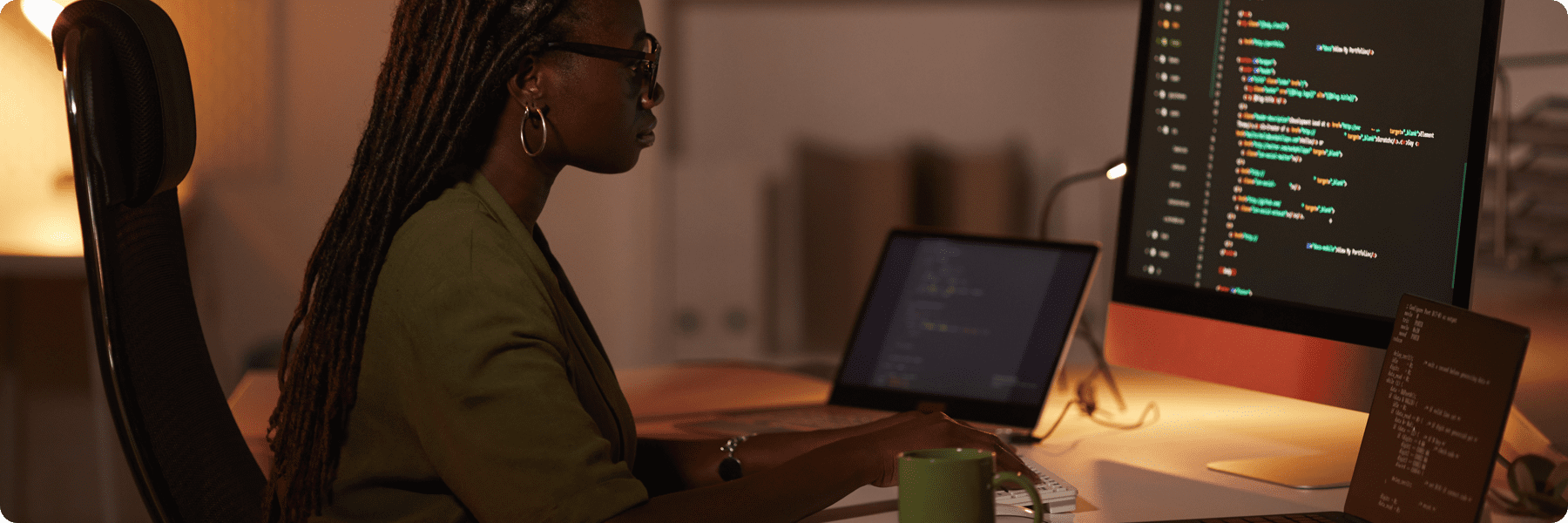 A person with long braided hair is seated at a desk in a digital marketing agency, typing on a computer. The screen showcases colorful code. A second monitor and a green mug adorn the desk. The room is dimly lit, fostering a focused atmosphere perfect for web design creativity.
