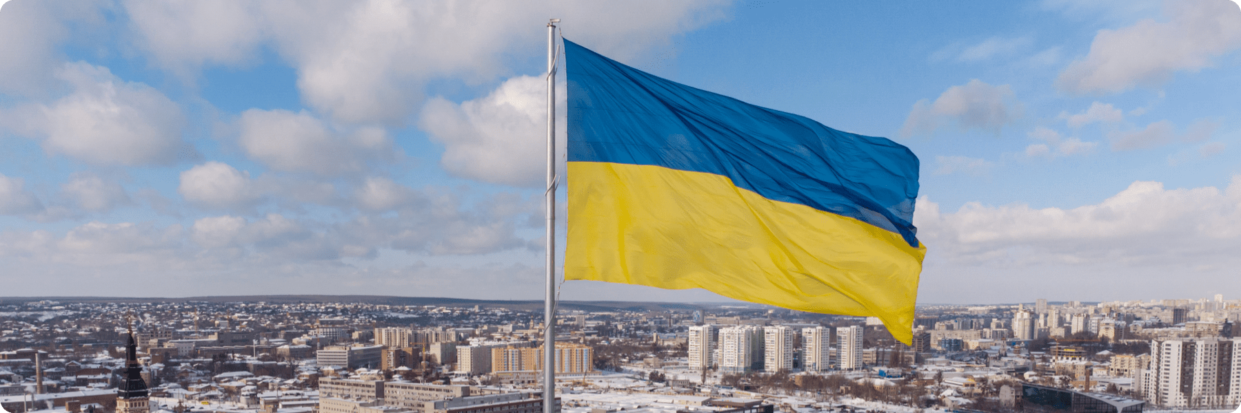 Large Ukrainian flag waving over a cityscape, perfectly captured by a digital marketing agency. Buildings and snow-covered streets are visible under a sky with scattered clouds, showcasing the art of web design in capturing the essence of Ukraines winter charm.