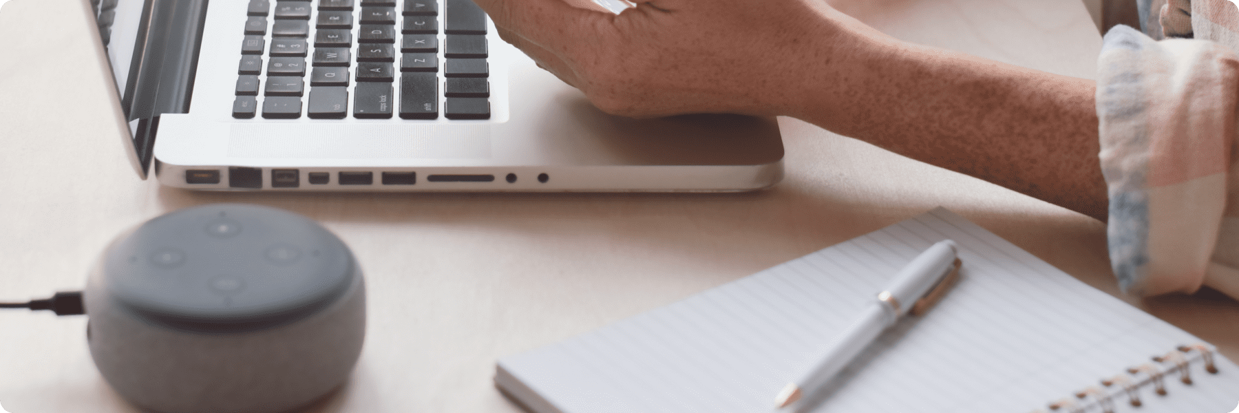 A person is immersed in digital marketing services, using a laptop and a smartphone. An open notebook with a pen rests on the right, while a smart speaker sits on the left, all elegantly arranged on a wooden table.