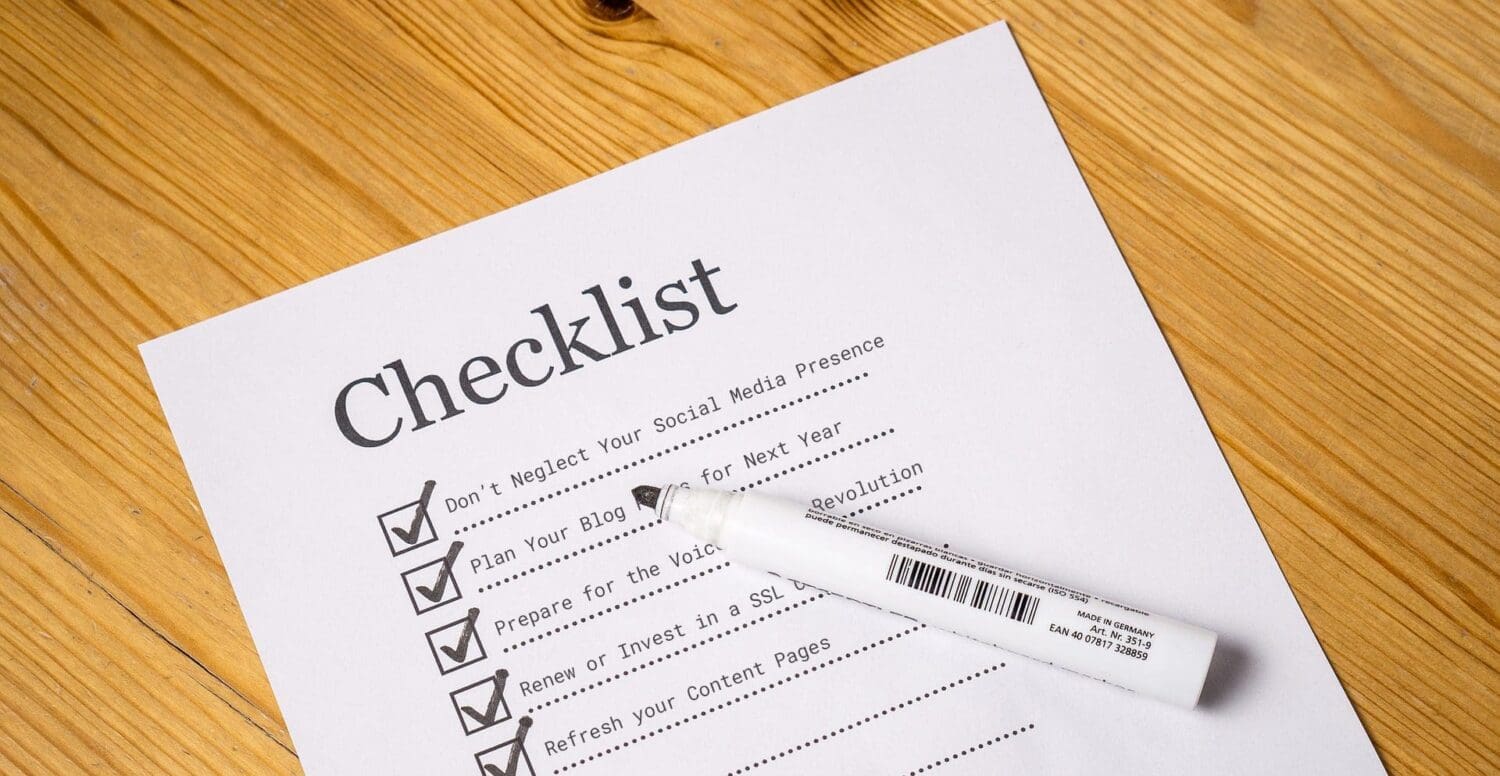 A printed checklist on a wooden table, partially checked off with a black marker lying next to it. Tasks include social media presence, blog planning, voice search preparation, SSL certificate renewal, content page refresh—all essential services offered by a digital marketing agency.