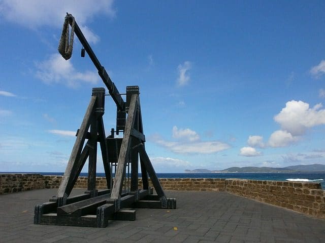 Perched on a stone platform overlooking the sea, the medieval trebuchet stands as tall and precise as a marketing agencys strategy, framed by a partly cloudy sky and distant mountains. Its wooden structure rises against the blue backdrop like an SEO service reaching new heights.