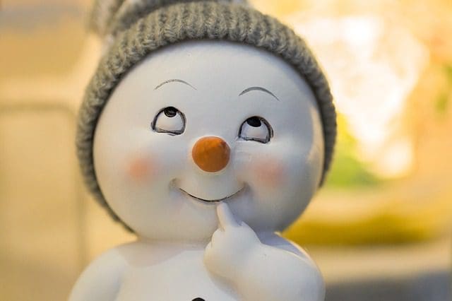 A ceramic snowman with a large orange nose and rosy cheeks dons a gray knitted hat. With an inquisitive expression, it gazes upward, finger on chin, pondering perhaps the next brilliant idea for a digital marketing company. The background is soft and blurred.