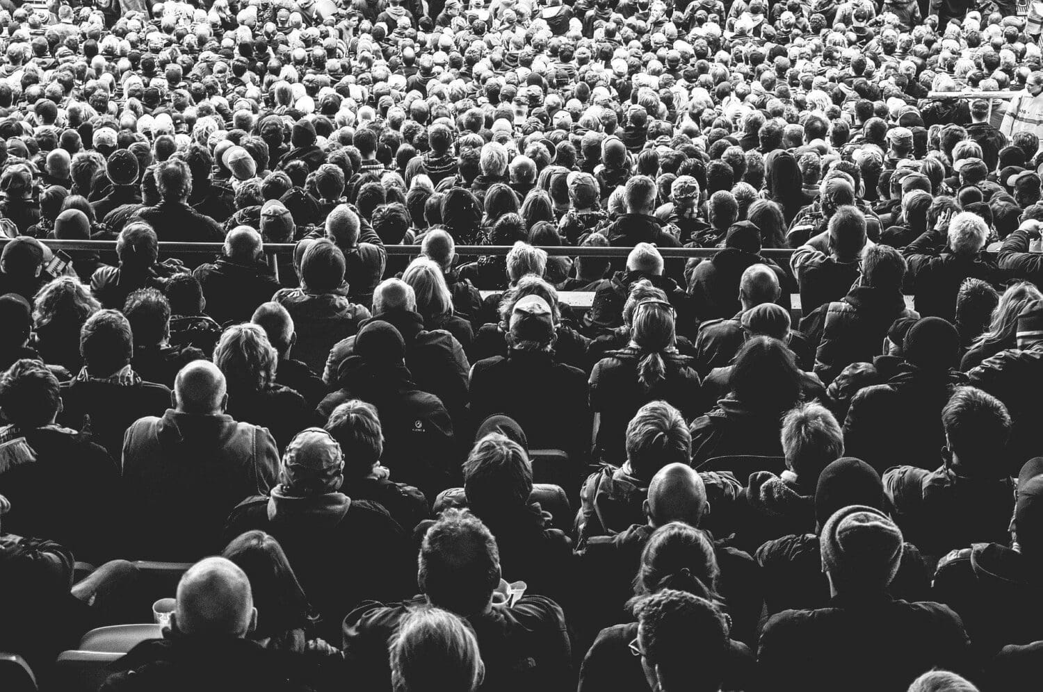 A black and white photo captures a large crowd seated in an auditorium, viewed from behind. The audience is tightly packed, each gaze fixed forward, much like attendees at a seminar by a digital marketing agency focusing on the latest in SEO services.