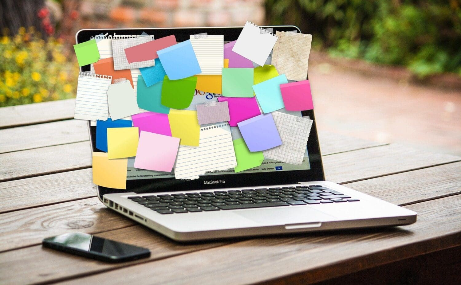 A laptop sits on a wooden table, its screen adorned with colorful sticky notes and paper sheets—perhaps reminders from a bustling digital marketing agency. Next to the laptop is a smartphone. In the background, plants lend a touch of nature to the scene.