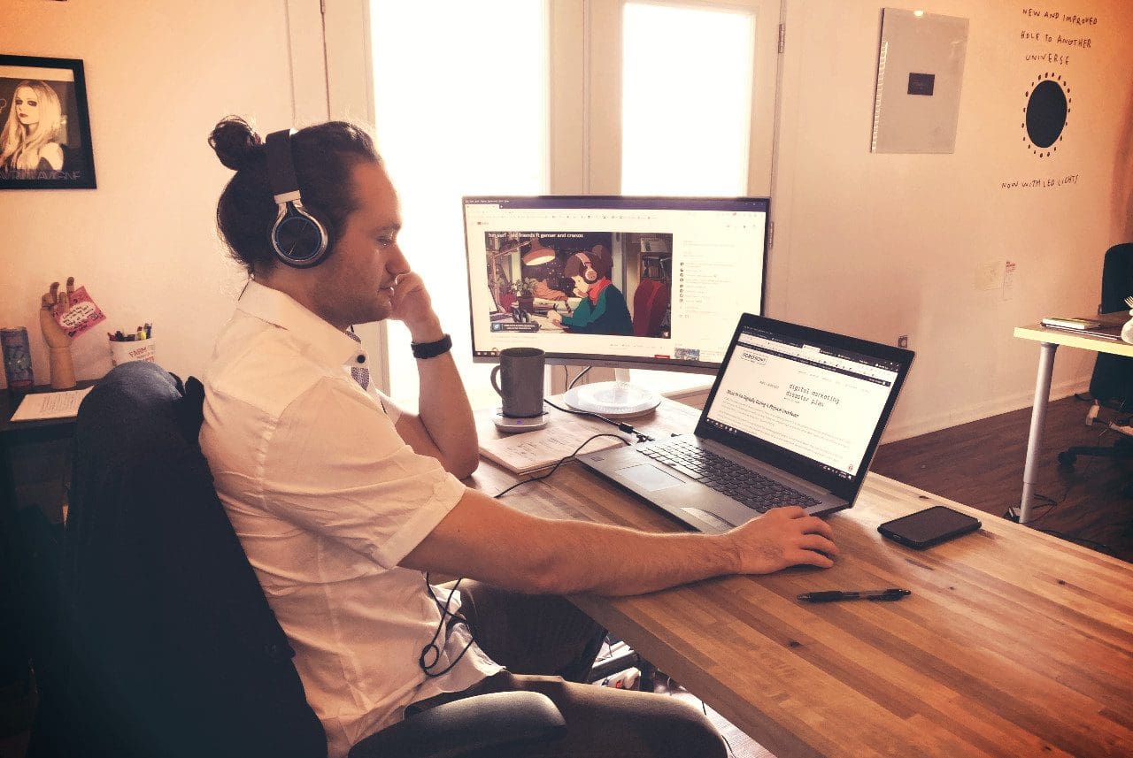 A person with headphones sits at a wooden desk, working on a laptop focused on web design. A monitor displays a video. The room is bright with sunlight, and a phone and coffee mug are on the desk. A framed poster and motivational wall art add inspiration to this creative workspace.