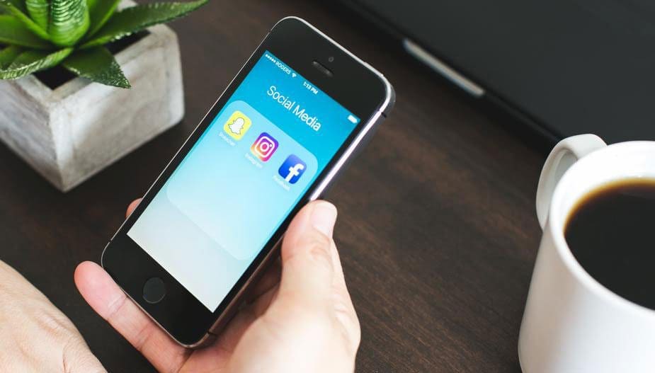 A person holds a smartphone displaying social media apps like Snapchat, Twitter, and Facebook. Beside the phone on the wooden table sits a potted plant and a cup of coffee, creating a perfect scene for any digital marketing company to envision new strategies.