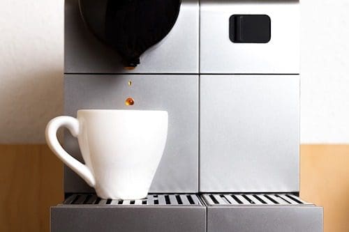 A sleek silver espresso machine dispenses coffee into a white mug on a metal grate tray, embodying the precision of web design. Two drops of coffee are suspended mid-air, offering a moment of perfection in focus while the background softly blurs.