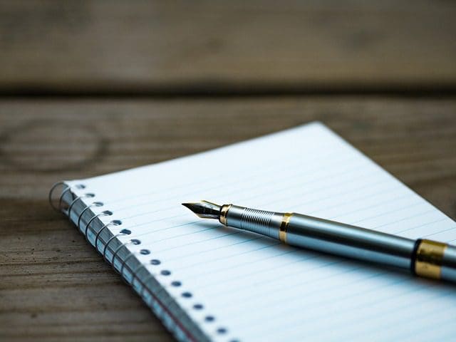 A fountain pen rests on top of an open spiral notebook with lined pages, looking like an analog tool in a digital marketing agency. The notebook is placed on a wooden surface, and the pen boasts a metallic finish with gold accents.