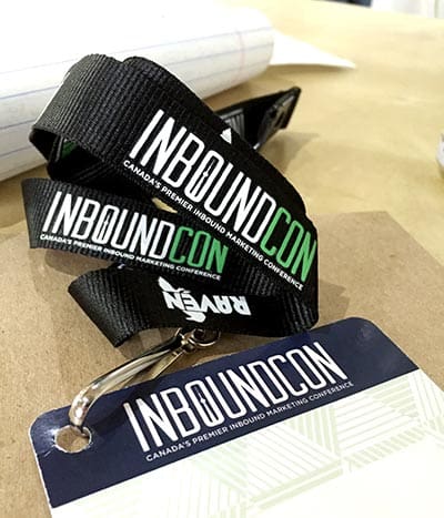 Close-up of a black lanyard with INBOUNDCON in white and green, alongside RAVEN in white. The attached conference badge features similar branding for Canadas premier inbound marketing conference, hosted by a leading digital marketing agency.