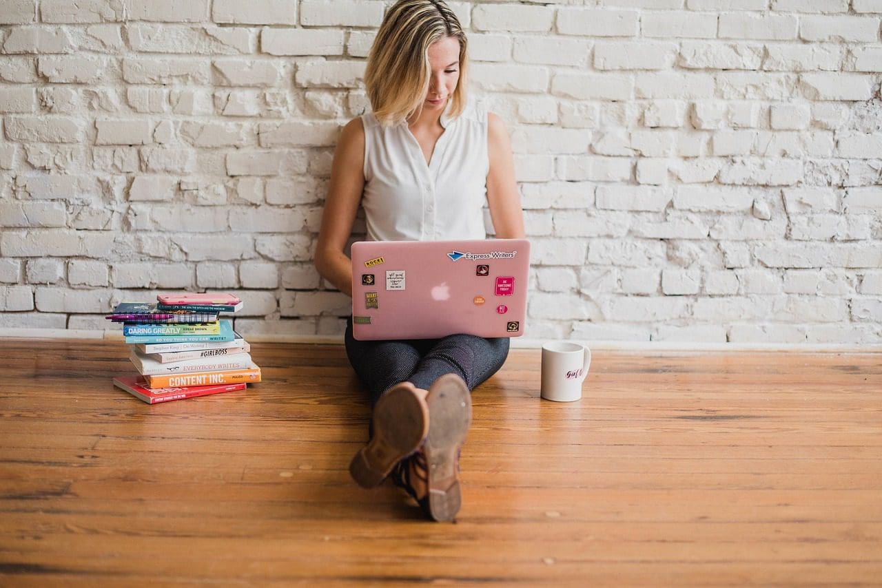 A person sits on a wooden floor leaning against a white brick wall, working on a pink laptop. A stack of books on their right hints at their focus on web design, while a white mug sits to their left. They wear a light sleeveless top and dark pants, immersed in digital creativity.