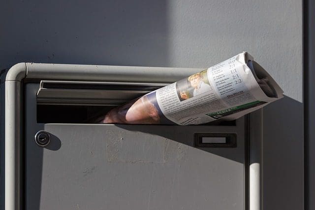 A rolled-up newspaper, like a message from the past, peeks out of a metal mailbox on a gray wall. Illuminated by sunlight, it casts shadows reminiscent of how SEO services can highlight content online. Sections of text and an image offer glimpses into stories to be discovered.