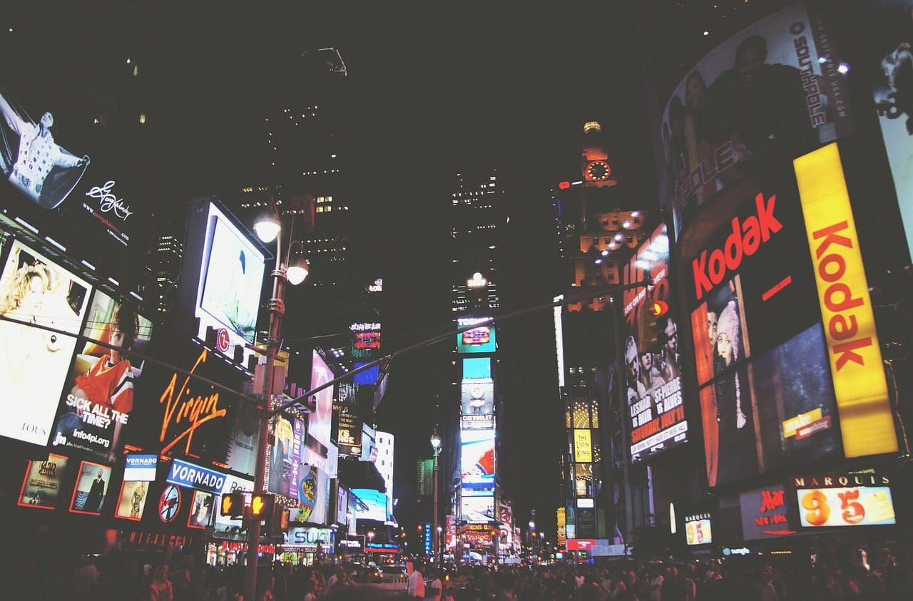 Night view of Times Square in New York City, bustling with crowds and illuminated by numerous bright, colorful advertisements and billboards. Prominent signs for Virgin and Kodak showcase the brilliance of digital marketing services. Skyscrapers loom in the background.