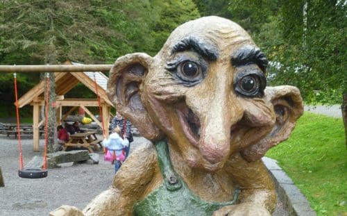 A large, whimsical troll statue with exaggerated features, including big ears and a long nose, stands near a playground with swings. Children and adults are visible in the background, surrounded by trees and picnic tables—a scene that could charm any digital marketing agency.