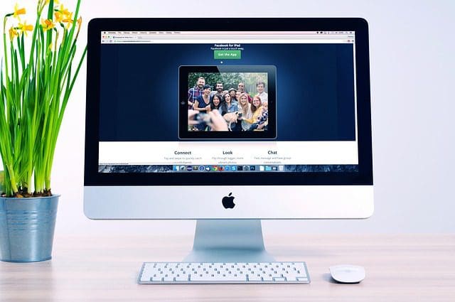An Apple iMac showcasing a social media page from a digital marketing agency, featuring a group photo. A vase with yellow flowers sits to the left of the computer, while a white mouse rests to the right on the desk.