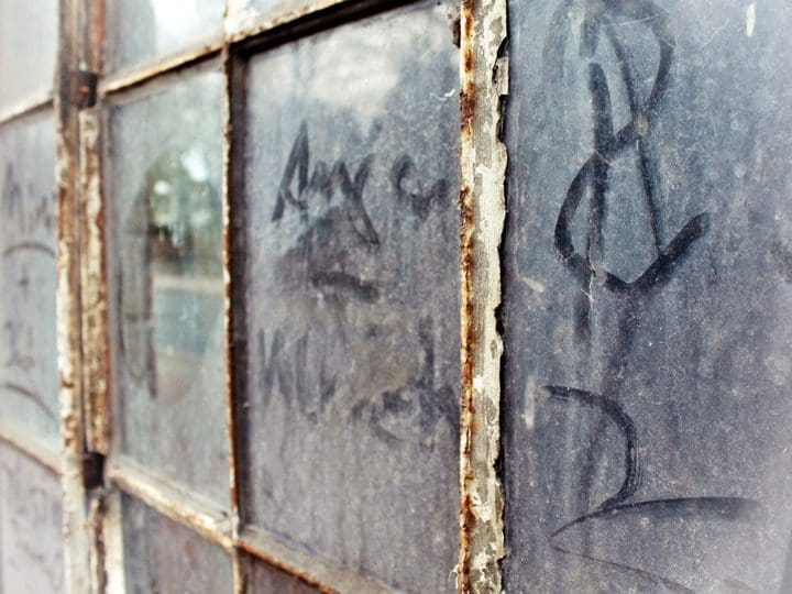 Close-up of a dusty, weathered window with cracked panes and rusted frames evokes the aging charm akin to an old digital marketing agency. Faint graffiti, including numbers and letters, adorns the glass surface while reflections of outdoor scenery linger in the background.