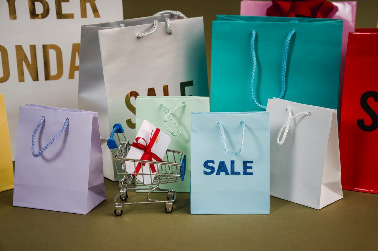 Colorful shopping bags with SALE text surround a small toy shopping cart holding a tiny white gift box with a red ribbon. In the background, partially visible words Cyber Monday hint at the digital marketing agency behind this eye-catching setup.