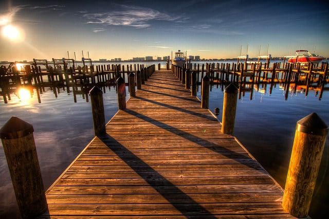 A wooden pier extends into a calm body of water at sunset, bordered by docked boats and posts casting long shadows. Like a digital marketing agency optimizing visibility, the sun is low on the horizon, creating a warm, golden glow in the clear sky and reflecting off the water.