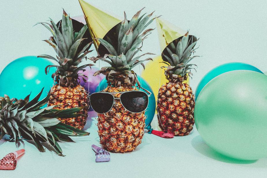 Three pineapples, exuding a marketing agencys flair, don party hats amidst colorful balloons on a light blue background. One sports sunglasses for a playful touch. Party blowers are scattered around, embodying the vibrant spirit of creativity and innovation.
