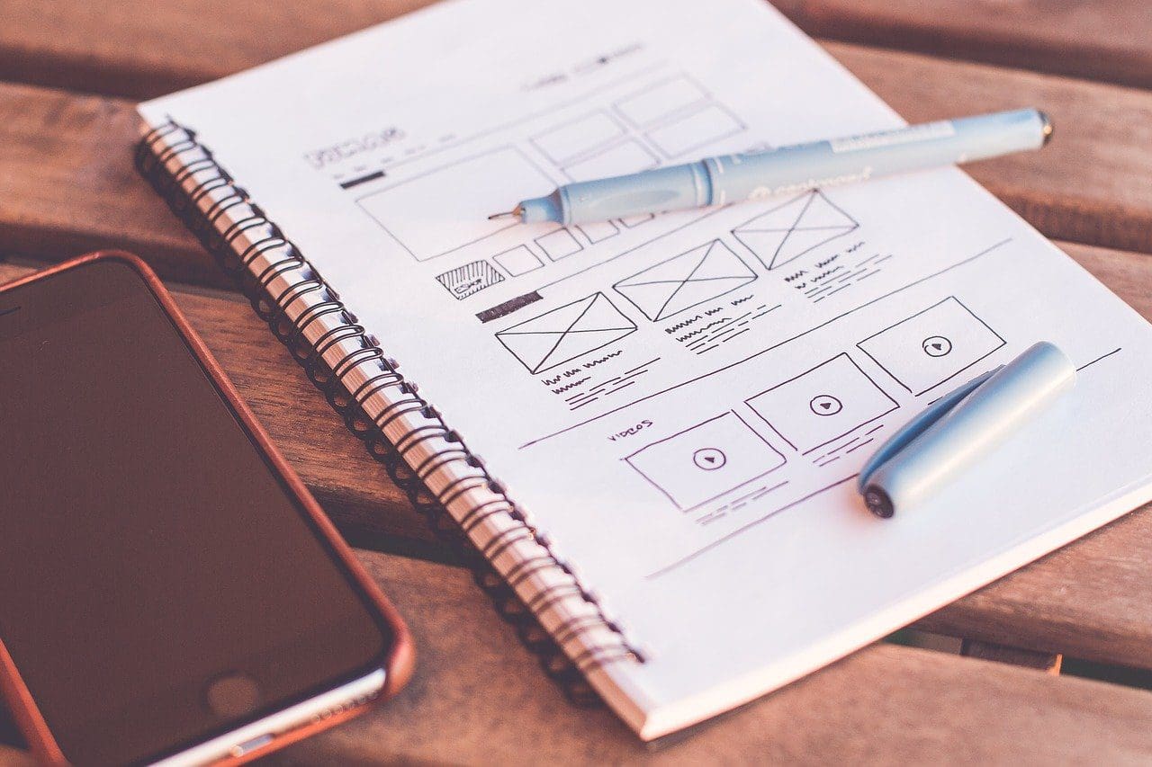A spiral notebook with website wireframe sketches lies on a wooden table, hinting at the planning phase for a digital marketing company. Two blue pens rest nearby, while a smartphone in a brown case completes this well-lit brainstorming scene.