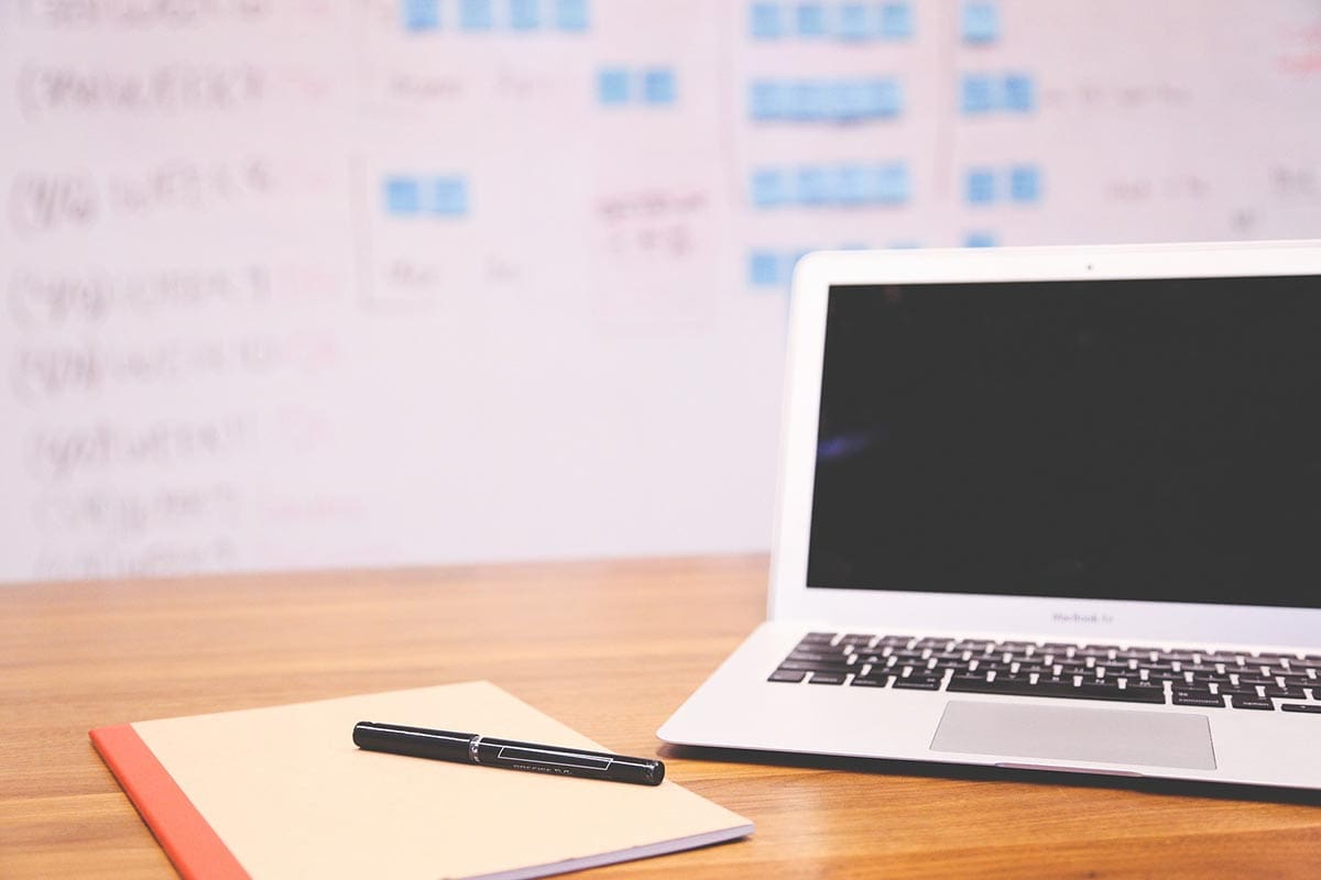 A laptop with a blank screen sits on a wooden desk beside a notebook and pen, ready for the next big idea. In the background, a whiteboard filled with notes on SEO services and web design strategies is slightly out of focus, capturing the essence of a digital marketing agency in action.
