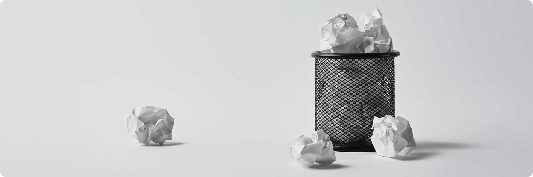 A black wire mesh trash can sits on a white surface, filled with crumpled paper balls. Three more lie scattered outside, echoing the chaos managed by a digital marketing agency. The scene remains minimalistic against a neutral background.