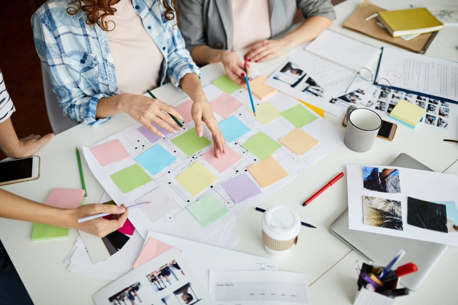 A group of people collaborating at a table covered with colorful sticky notes, papers, and design layouts at a digital marketing agency. They are holding pens and discussing ideas. A coffee cup and smartphone are also visible on the table, hinting at SEO services in progress.