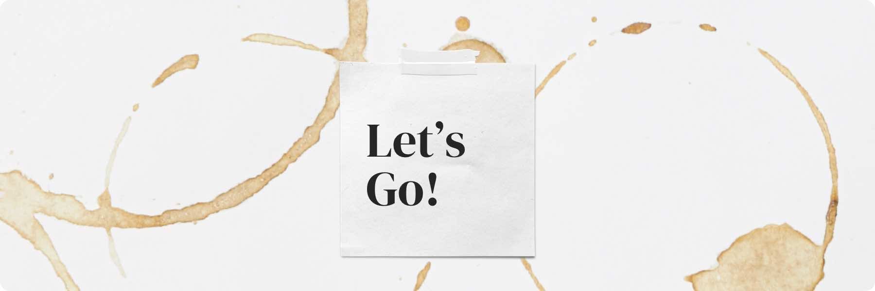 A piece of paper with the text Lets Go! is taped to a white background adorned with coffee cup ring stains—a quirky reminder of brainstorms at our digital marketing agency.