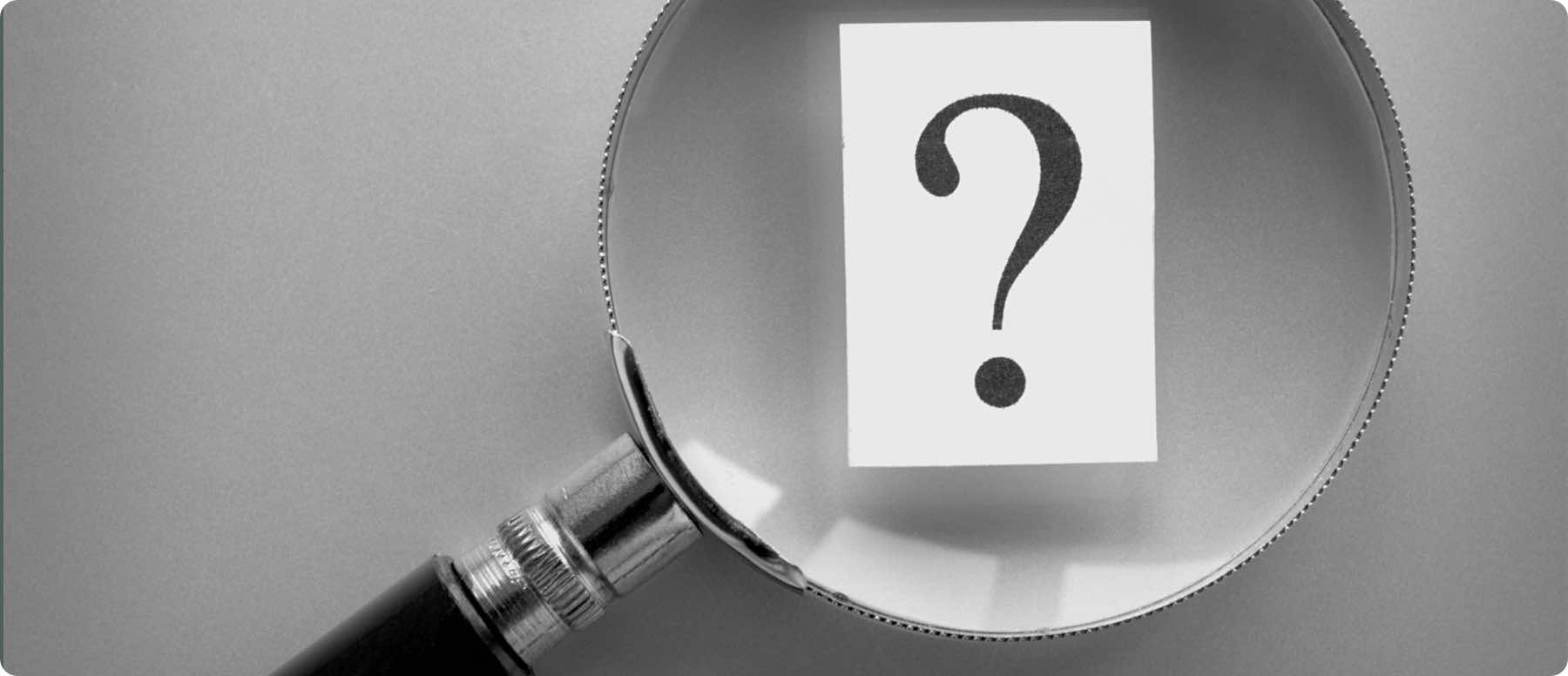 A magnifying glass enlarges a question mark on a small piece of paper, set against a grayscale backdrop. This scene symbolizes curiosity and inquiry—key traits for any digital marketing company seeking innovative solutions.