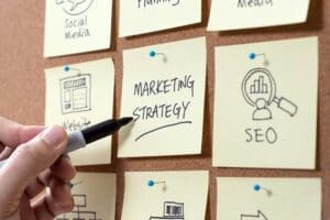 A hand scribbles Marketing Strategy on a yellow sticky note pinned to a corkboard. Nearby, notes with hand-drawn icons and labels like Website, Social Media, and SEO services hint at a digital marketing companys brainstorming session.