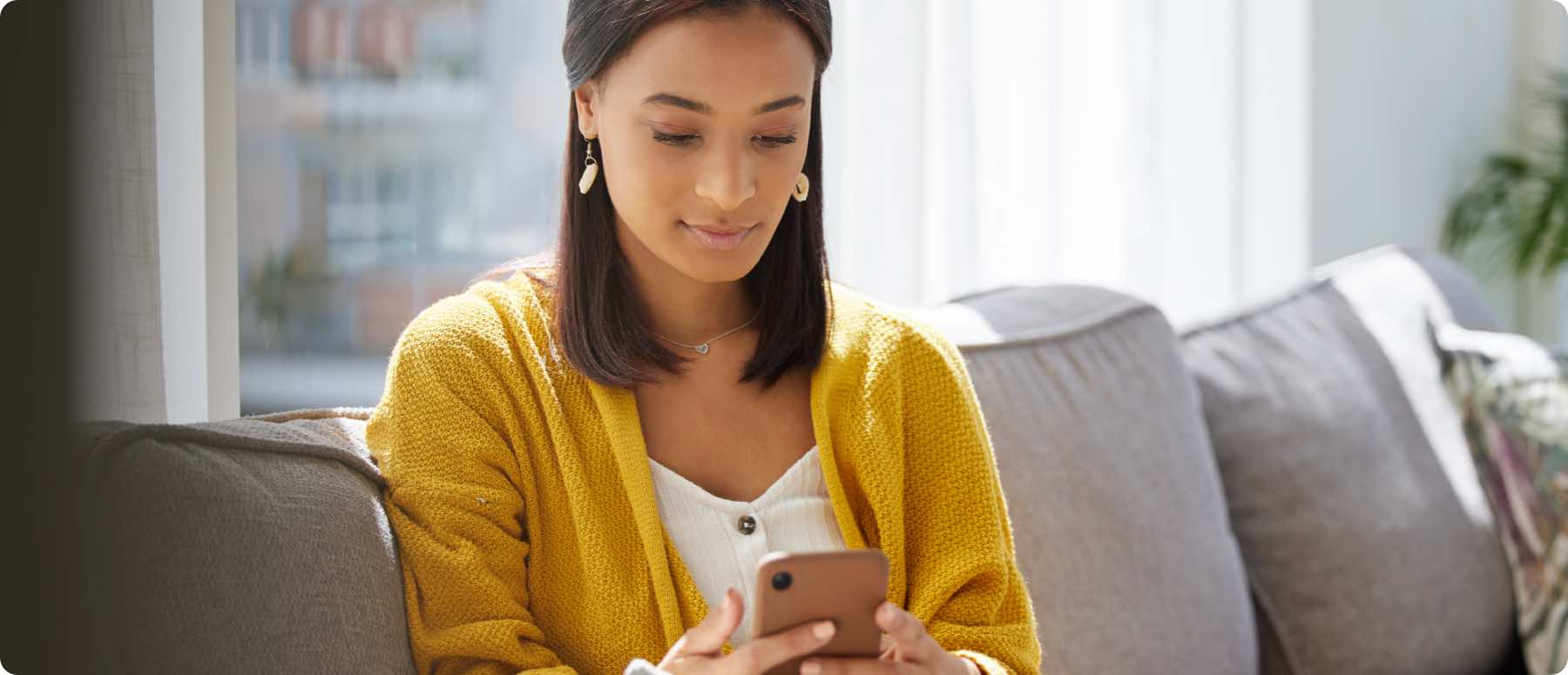 A woman sits on a gray sofa in a bright room, looking at her smartphone, perhaps browsing digital marketing services. She is wearing a yellow cardigan and white top, with earrings and a necklace. Sunlight streams through a window, illuminating the scene.