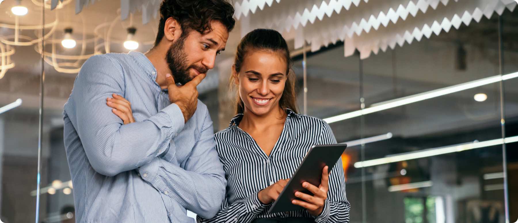 A man and woman stand in an office, looking at a tablet together. The woman, smiling, points at the tablet displaying digital marketing services. The man looks thoughtful with his hand on his chin. Both are dressed in business attire.