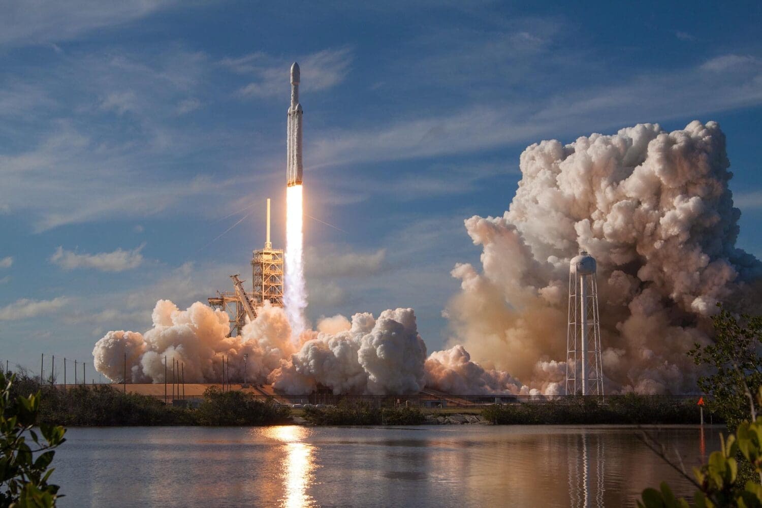 A rocket is launching into a blue sky, leaving a plume of smoke behind. The scene, reminiscent of digital marketing agency creativity, is reflected in a body of water in the foreground, with vegetation on the lower left.