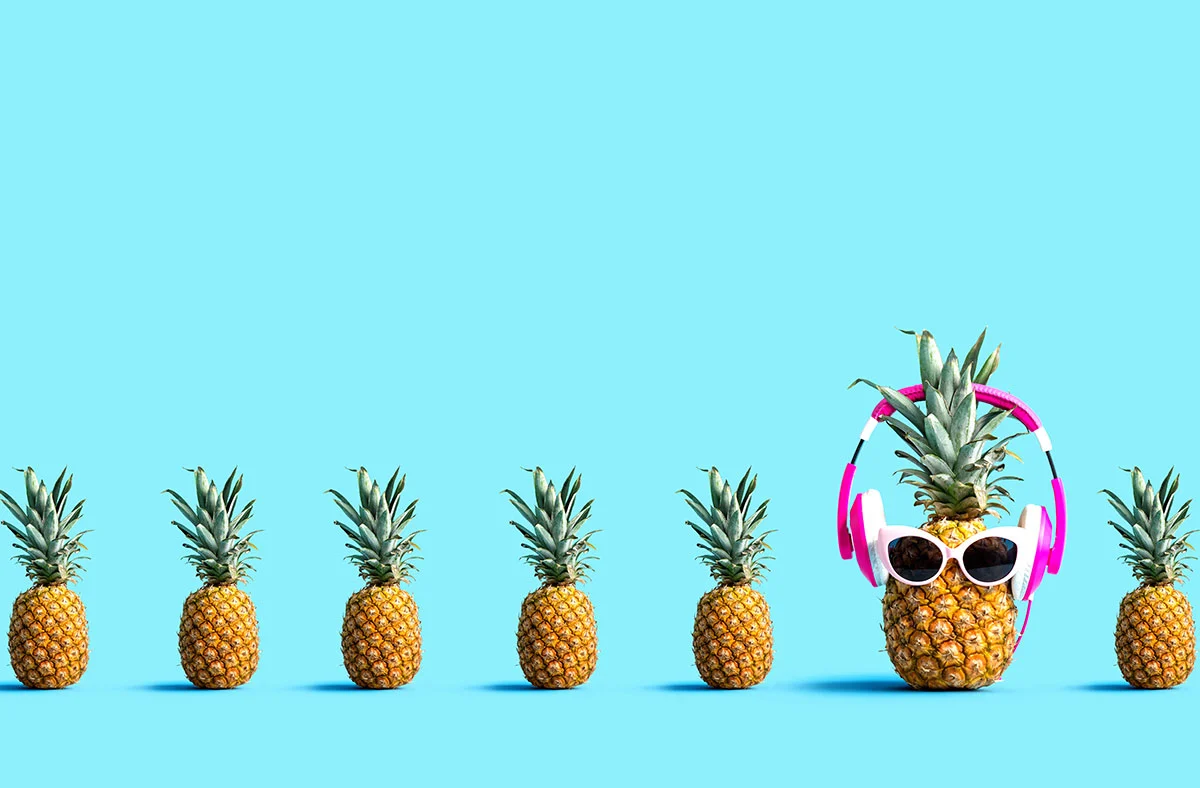Five pineapples are lined up on a blue background. The last one is larger, adorned with pink headphones and sunglasses, creating a playful contrast—much like how a creative digital marketing agency stands out in web design.