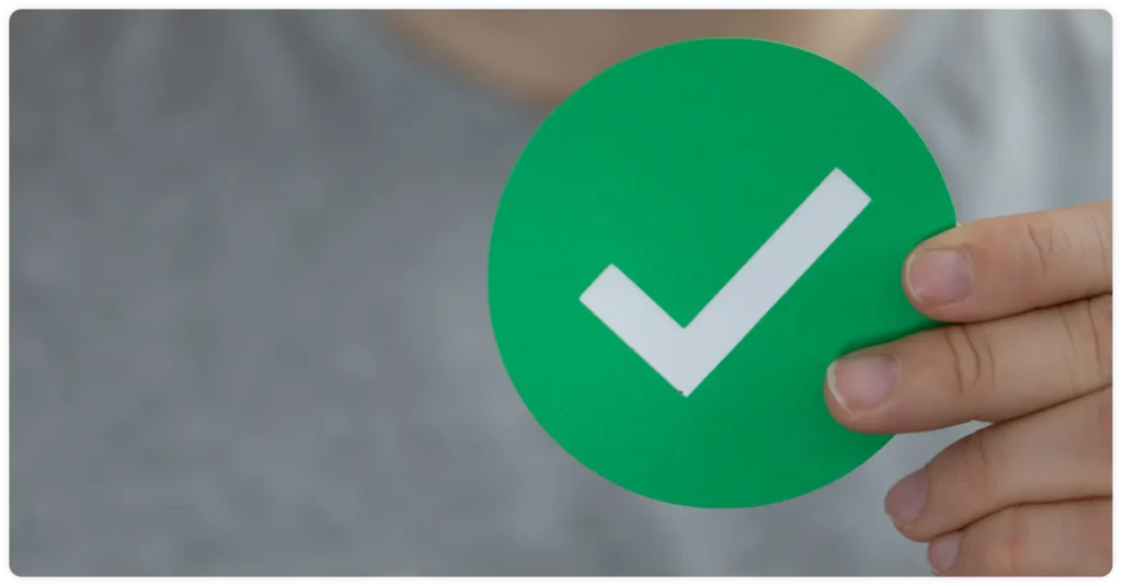 A person holds a green circle with a large white check mark on it, symbolizing approval or completion. The background is out of focus.