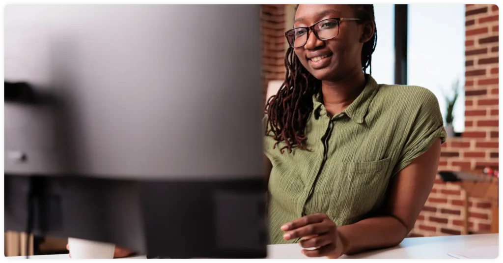 A woman wearing glasses and a green shirt sits at a desk, smiling while working on a computer in a modern office with brick walls.