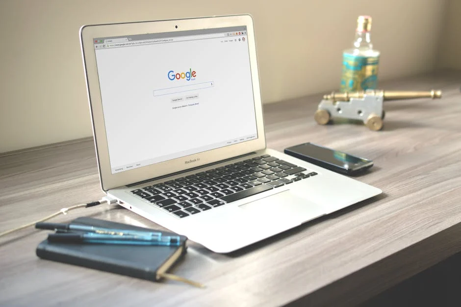 A silver laptop displaying the Google homepage sits on a wooden desk beside a smartphone, a closed notebook with a pen, and a decorative bottle next to a small brass cannon ornament.