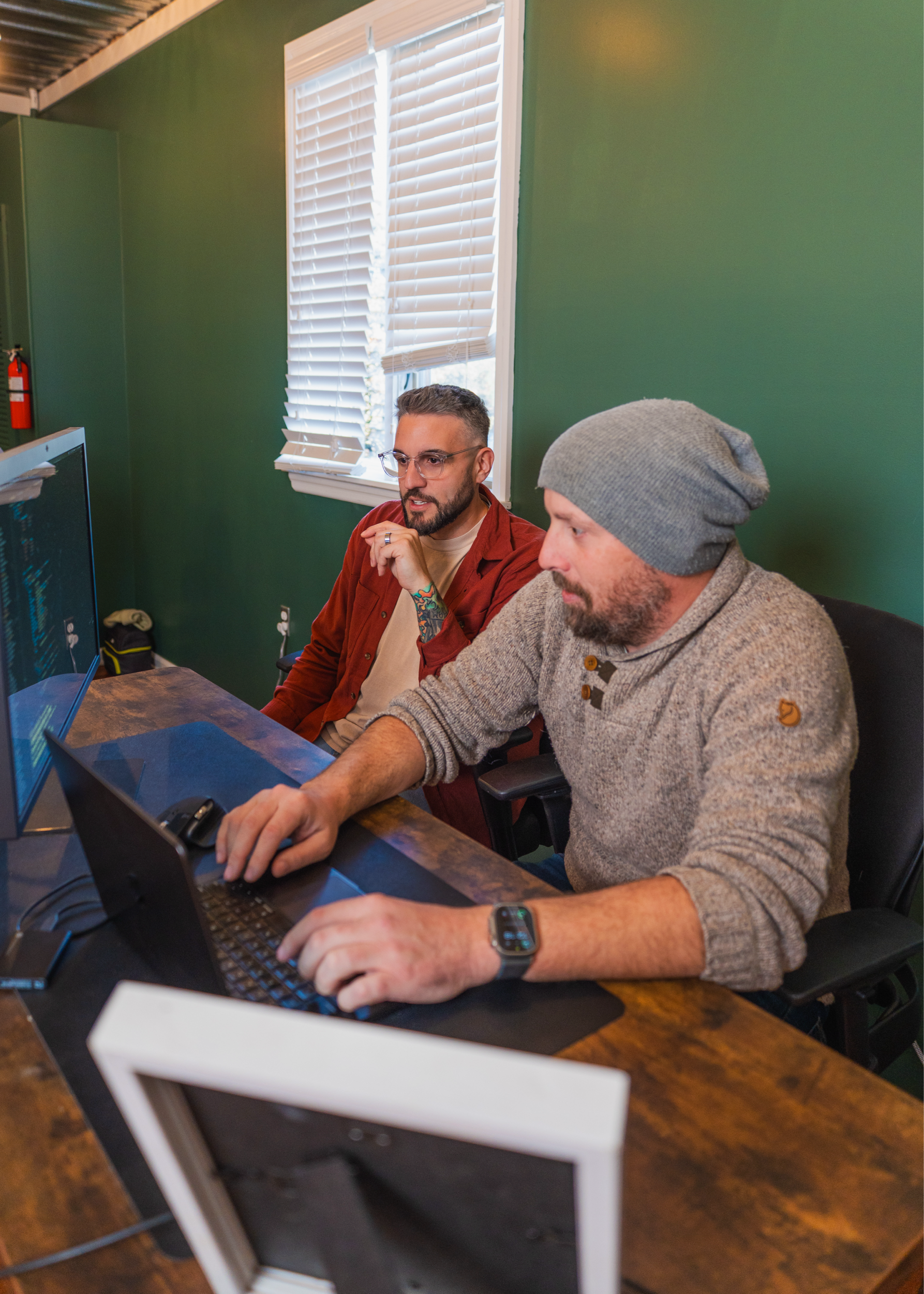 Two men sit at a wooden desk in front of computer monitors in a room with green walls and a window with blinds. One man types on a laptop while the other looks at the screen thoughtfully.
